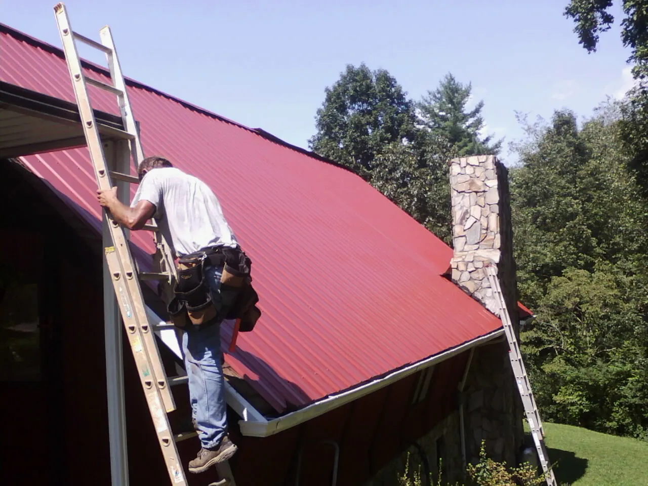 Roofer on a ladder beside a red metal roof and stone chimney — Soffit & Fascia Repair by Roofing Brothers in Greenwich